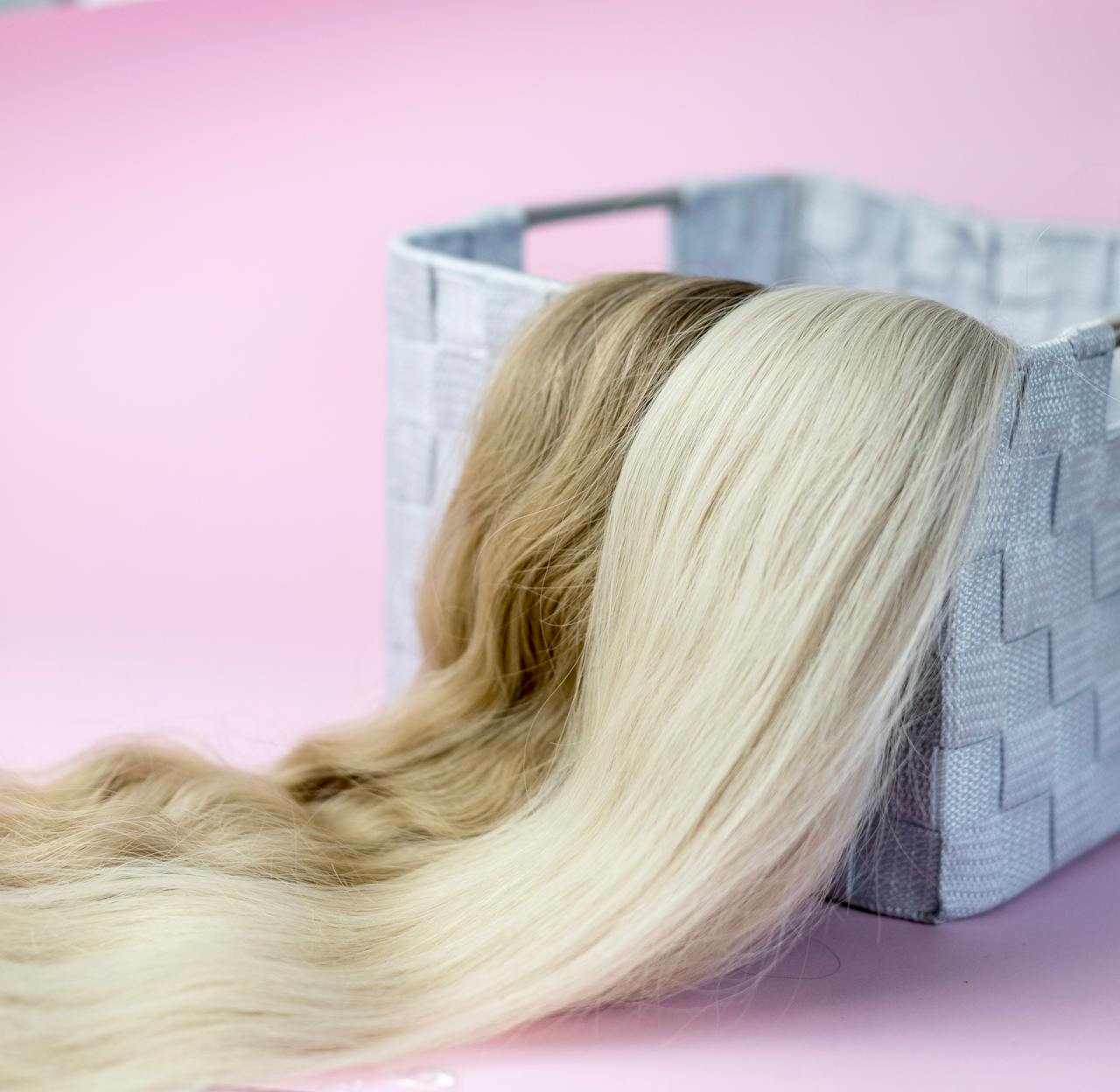 programs-03 Close-up of blond and brown hair extensions draped over a woven basket, on a pink background.